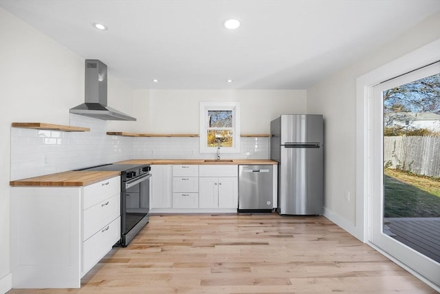 kitchen with wall chimney range hood, sink, butcher block countertops, white cabinetry, and stainless steel appliances