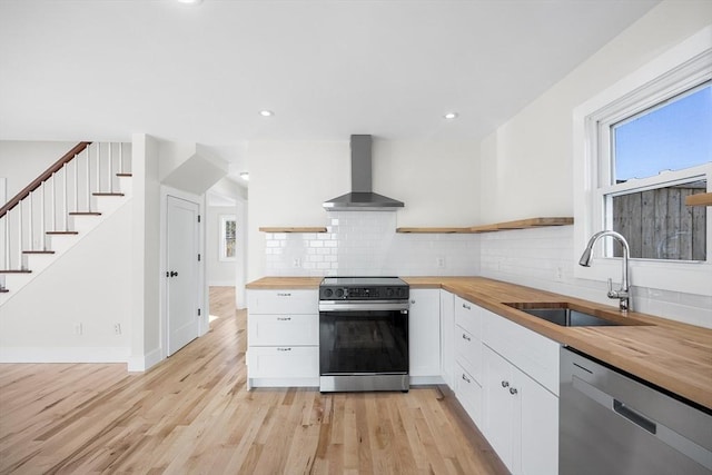 kitchen with sink, stainless steel appliances, wall chimney range hood, wooden counters, and white cabinets