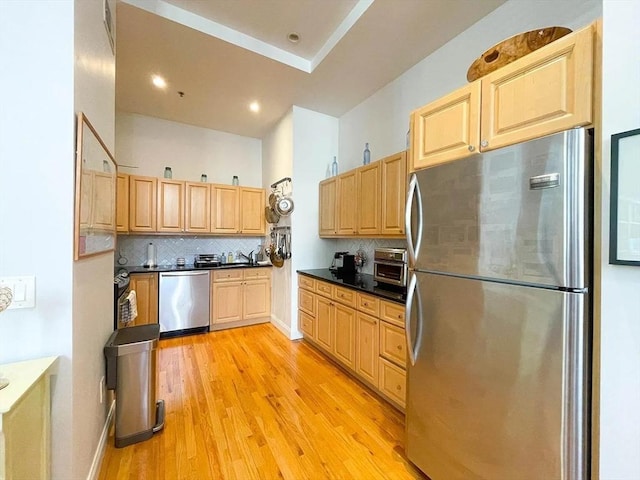 kitchen featuring appliances with stainless steel finishes, tasteful backsplash, light brown cabinets, light wood-style floors, and dark countertops