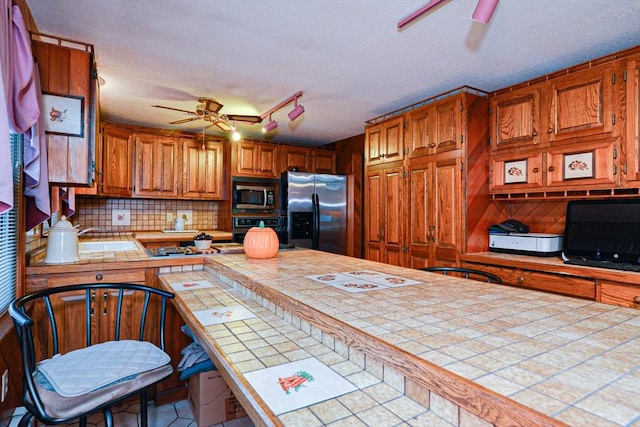 kitchen with stainless steel appliances, a ceiling fan, tile countertops, decorative backsplash, and brown cabinetry