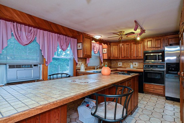 kitchen with appliances with stainless steel finishes, tile countertops, a breakfast bar area, backsplash, and brown cabinetry