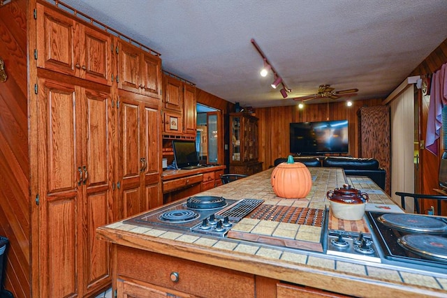 kitchen with tile countertops, a ceiling fan, brown cabinetry, a textured ceiling, and wooden walls