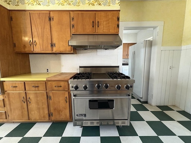 kitchen featuring brown cabinetry, light floors, luxury stove, freestanding refrigerator, and under cabinet range hood