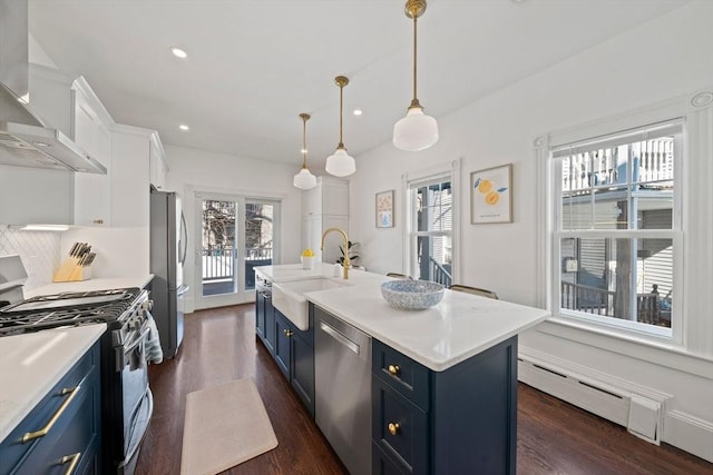 kitchen with appliances with stainless steel finishes, blue cabinets, baseboard heating, wall chimney range hood, and a sink