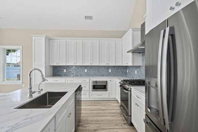 kitchen with a sink, white cabinetry, light wood-style floors, appliances with stainless steel finishes, and wall chimney range hood