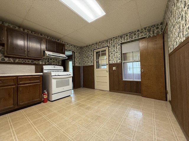 kitchen with white range with gas stovetop, under cabinet range hood, a wainscoted wall, wallpapered walls, and dark brown cabinets