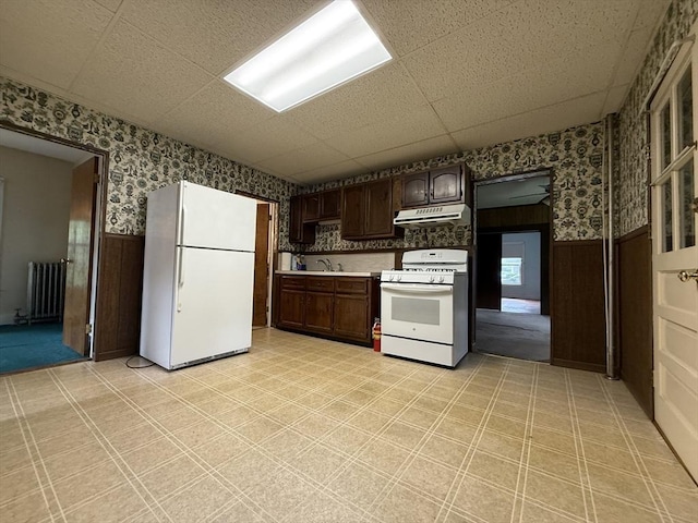 kitchen with white appliances, under cabinet range hood, a wainscoted wall, radiator heating unit, and wallpapered walls