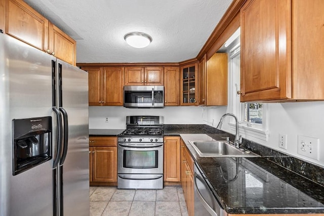 kitchen with dark stone countertops, glass insert cabinets, appliances with stainless steel finishes, brown cabinets, and a sink