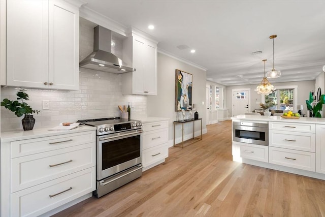 kitchen featuring light wood-type flooring, stainless steel appliances, crown molding, wall chimney range hood, and white cabinetry