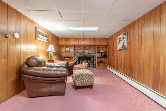 carpeted living room featuring a warm lit fireplace, wooden walls, baseboard heating, and a drop ceiling