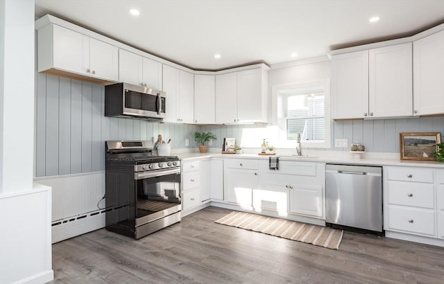 kitchen with stainless steel appliances, baseboard heating, white cabinetry, a sink, and wood finished floors