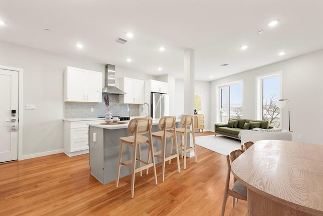 kitchen featuring wall chimney range hood, light wood-style floors, freestanding refrigerator, white cabinetry, and decorative backsplash