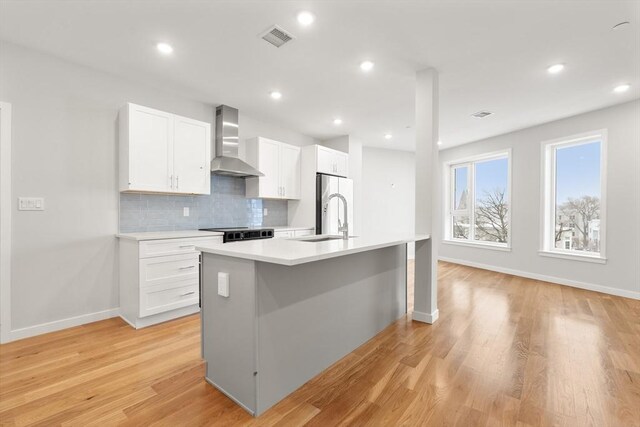 kitchen featuring visible vents, an island with sink, wall chimney range hood, light countertops, and a sink