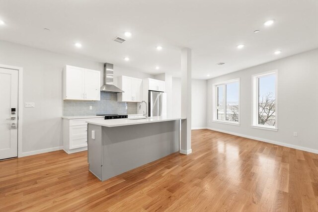 kitchen with light countertops, white cabinets, freestanding refrigerator, wall chimney exhaust hood, and an island with sink