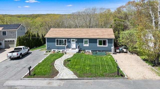 single story home featuring asphalt driveway, a front yard, a forest view, and roof with shingles