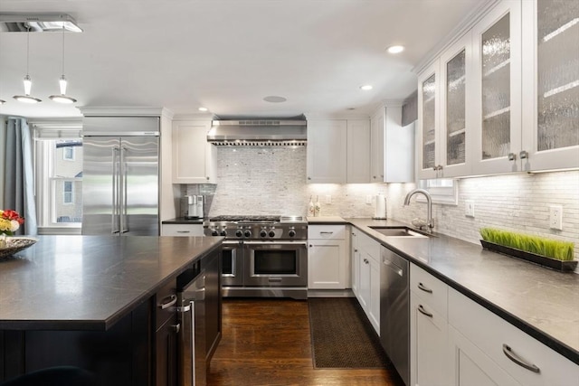 kitchen with a sink, high end appliances, wall chimney exhaust hood, dark countertops, and white cabinetry