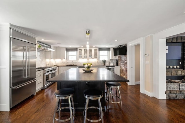 kitchen with high end appliances, dark countertops, a sink, wall chimney exhaust hood, and a breakfast bar area