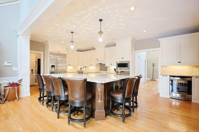 kitchen with decorative light fixtures, a center island with sink, white cabinets, built in appliances, and beverage cooler