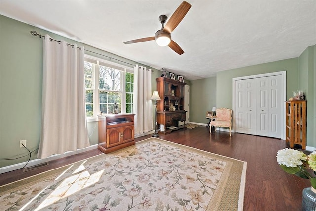 living area with ceiling fan, visible vents, a wood stove, wood finished floors, and baseboards