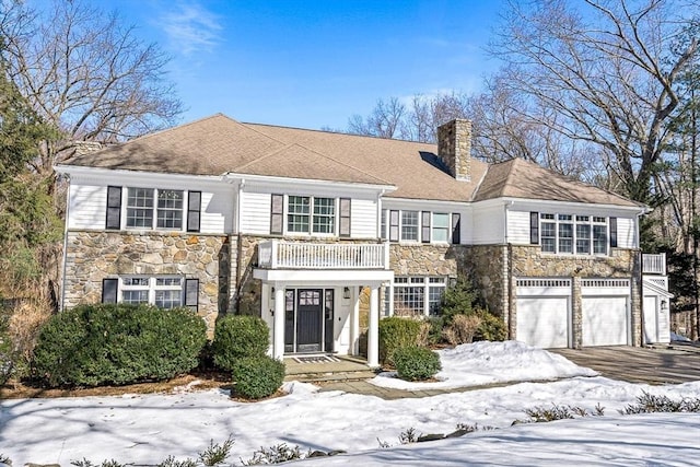 view of front of house with a balcony, a garage, stone siding, driveway, and a chimney