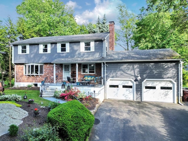 colonial inspired home featuring aphalt driveway, covered porch, a garage, brick siding, and a chimney