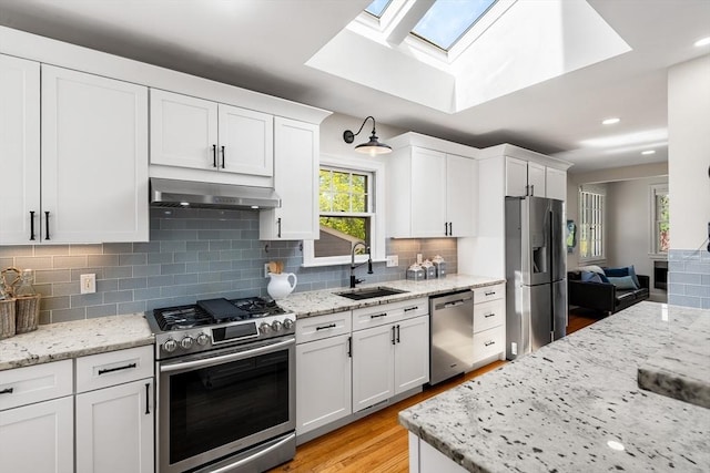 kitchen featuring white cabinetry, under cabinet range hood, a sink, appliances with stainless steel finishes, and a skylight