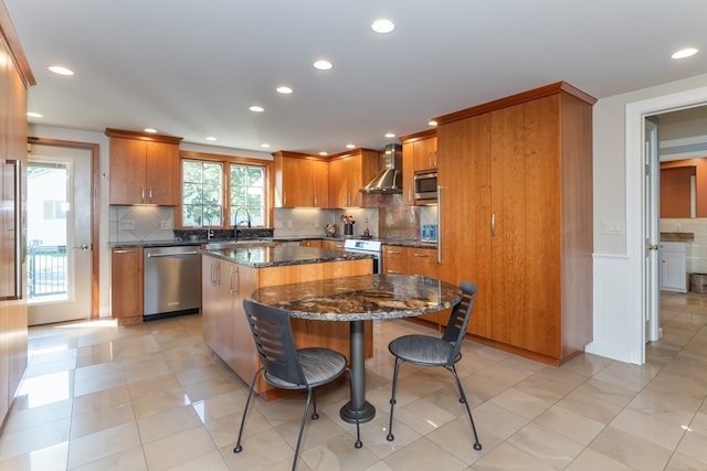 kitchen with dark stone counters, a kitchen bar, a center island, wall chimney exhaust hood, and appliances with stainless steel finishes