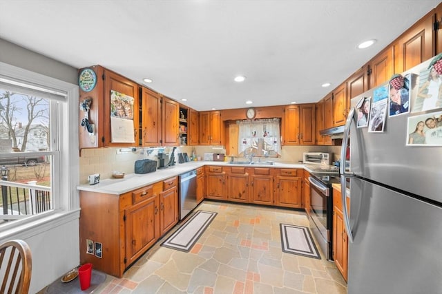 kitchen featuring under cabinet range hood, stainless steel appliances, a sink, brown cabinets, and stone finish flooring