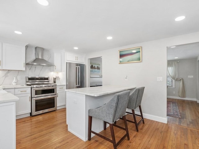 kitchen featuring backsplash, white cabinets, a kitchen breakfast bar, stainless steel appliances, and wall chimney range hood