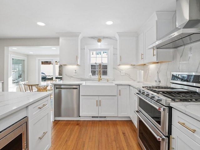 kitchen with sink, white cabinetry, stainless steel appliances, light stone countertops, and wall chimney exhaust hood