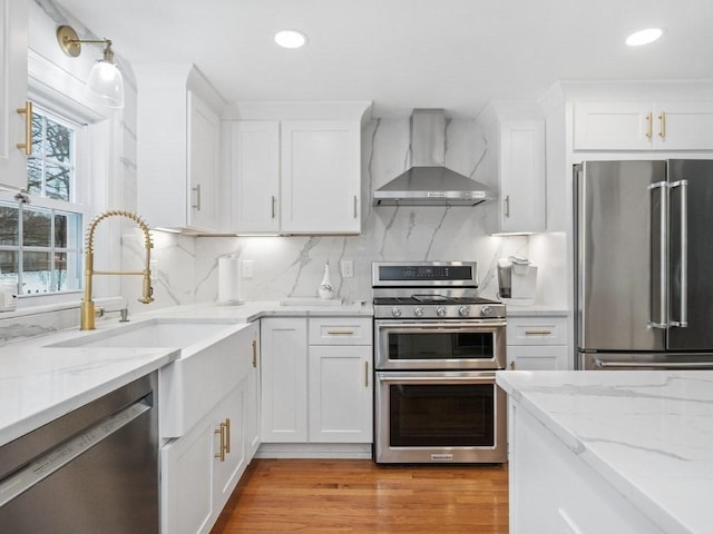 kitchen with white cabinetry, stainless steel appliances, light stone counters, and wall chimney range hood