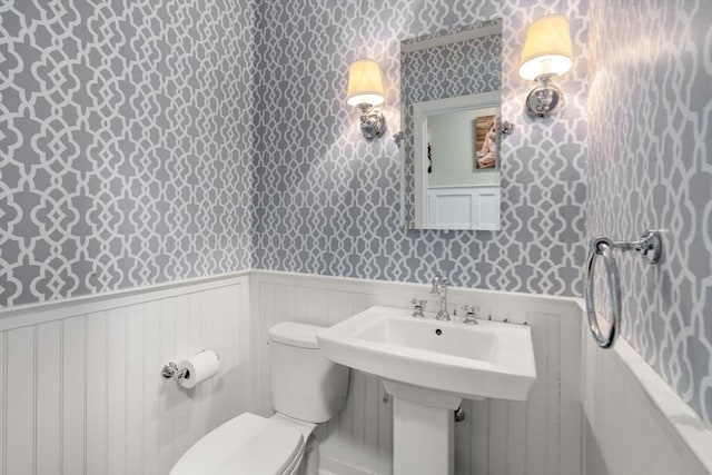 half bath with wallpapered walls, toilet, and wainscoting
