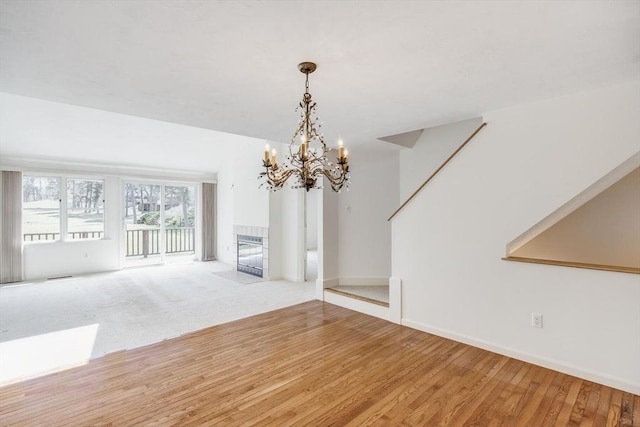 unfurnished living room with a notable chandelier, a tiled fireplace, baseboards, wood finished floors, and stairway