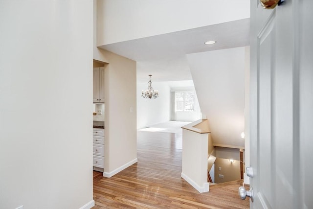 corridor featuring baseboards, a notable chandelier, light wood-type flooring, and an upstairs landing