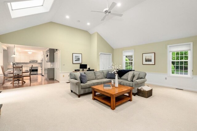 living area featuring high vaulted ceiling, ceiling fan, a skylight, a wainscoted wall, and light carpet
