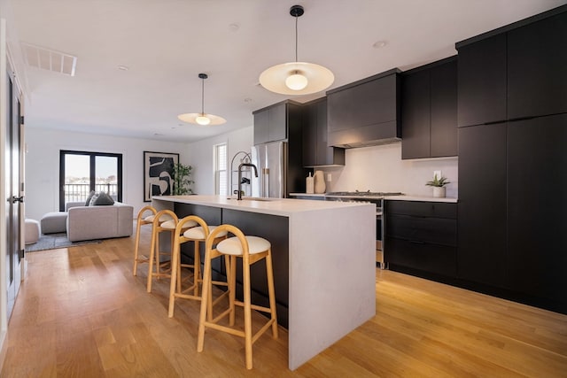 kitchen with custom exhaust hood, stainless steel appliances, a kitchen island with sink, light hardwood / wood-style flooring, and hanging light fixtures