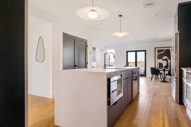 kitchen featuring pendant lighting, stainless steel oven, a center island with sink, sink, and light wood-type flooring