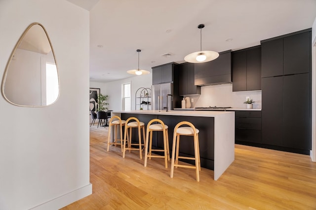 kitchen featuring pendant lighting, premium range hood, a kitchen island with sink, light hardwood / wood-style floors, and stainless steel appliances