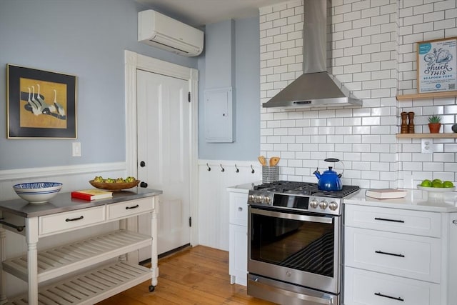 kitchen featuring stainless steel range with gas cooktop, electric panel, white cabinets, a wall mounted air conditioner, and wall chimney range hood