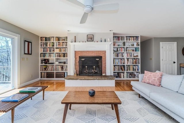 living area featuring light wood finished floors, a brick fireplace, built in features, a ceiling fan, and baseboards