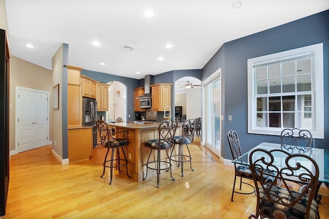 kitchen featuring stainless steel appliances, stone countertops, a breakfast bar, light hardwood / wood-style flooring, and wall chimney exhaust hood