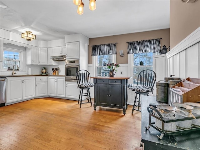 kitchen featuring under cabinet range hood, stainless steel appliances, white cabinetry, light wood-style floors, and wainscoting