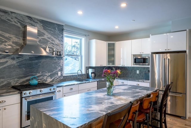 kitchen with a kitchen island, stainless steel appliances, wall chimney range hood, white cabinetry, and a sink