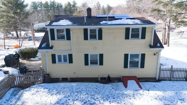 snow covered back of property with fence, a chimney, and solar panels