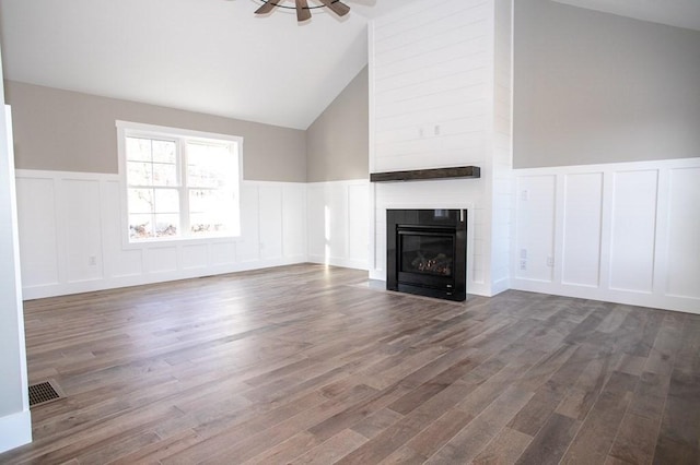 unfurnished living room featuring a decorative wall, a large fireplace, a wainscoted wall, wood finished floors, and a ceiling fan