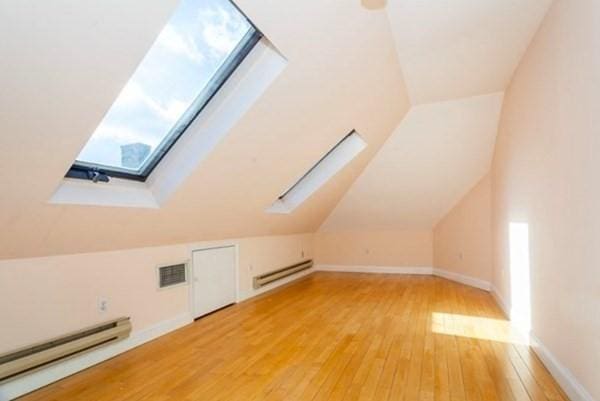 additional living space featuring vaulted ceiling with skylight, a wealth of natural light, light wood finished floors, and baseboard heating