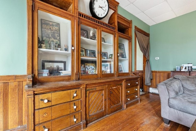 interior space featuring wainscoting, baseboard heating, wood walls, a paneled ceiling, and dark wood-type flooring