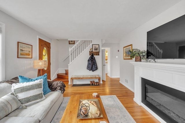 living room featuring a glass covered fireplace, light wood-type flooring, stairs, and baseboards