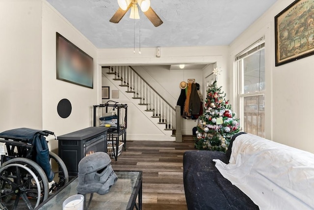 living area with ceiling fan, a wood stove, baseboards, dark wood-style floors, and stairs