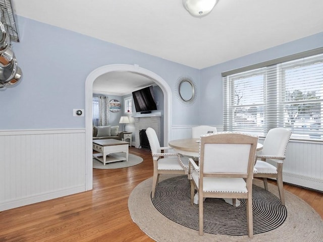 dining room with arched walkways, light wood finished floors, wainscoting, and a brick fireplace